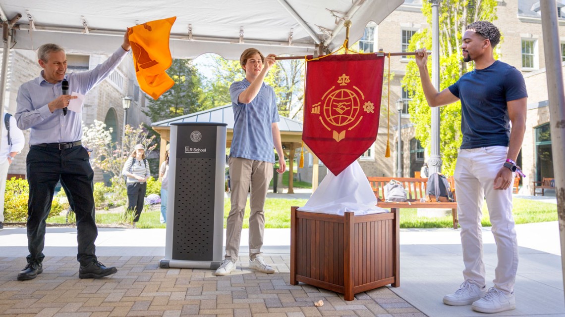Alex Colvin, Ph.D. '99, the Kenneth F. Kahn ’69 Dean and the Martin F. Scheinman ’75, MS ’76, Professor of Conflict Resolution, unveils the new ceremonial banner at an event held May 7. Holding the updated flag are Patrick Raczka ’25 (left) and Tyler Bonaparte ’25 (right), who were part of the student team that created the winning redesign.