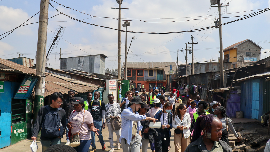 Students walk with local community members down a street between homes and shops