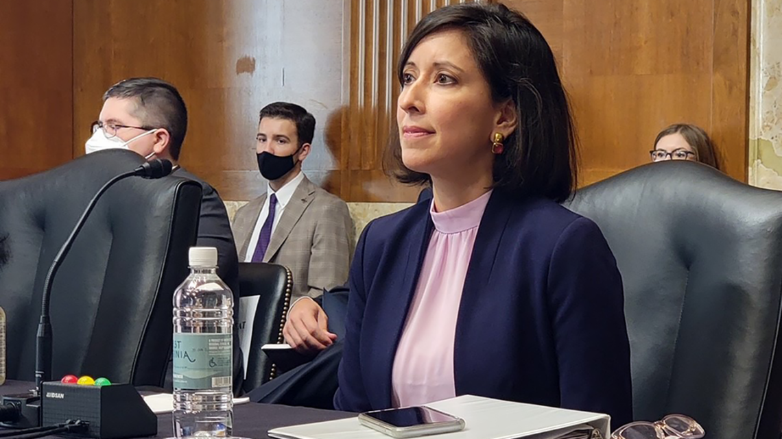 Woman in a blue suit seated at a table in front of a microphone with spectators gathered behind her.